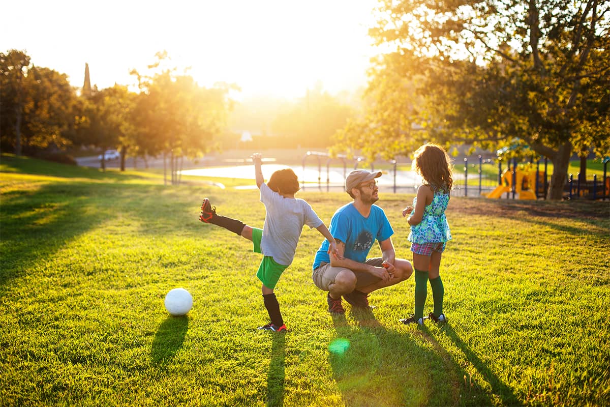 Man talking to child in park as another child kicks a ball