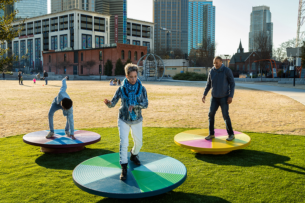 two adults and a child playing on playground equipment in front of the Charlotte, NC skyline