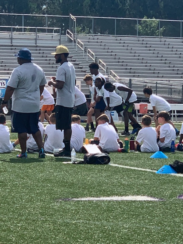 children sitting on a football field while other children prepare for a running race