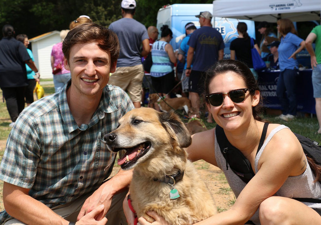 Two people smiling with a dog with a group of people behind them